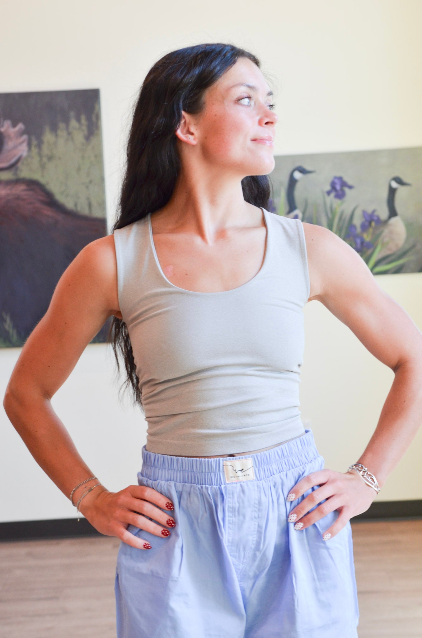 Woman wearing a light gray tank top and light blue pants in a room with paintings on the wall.