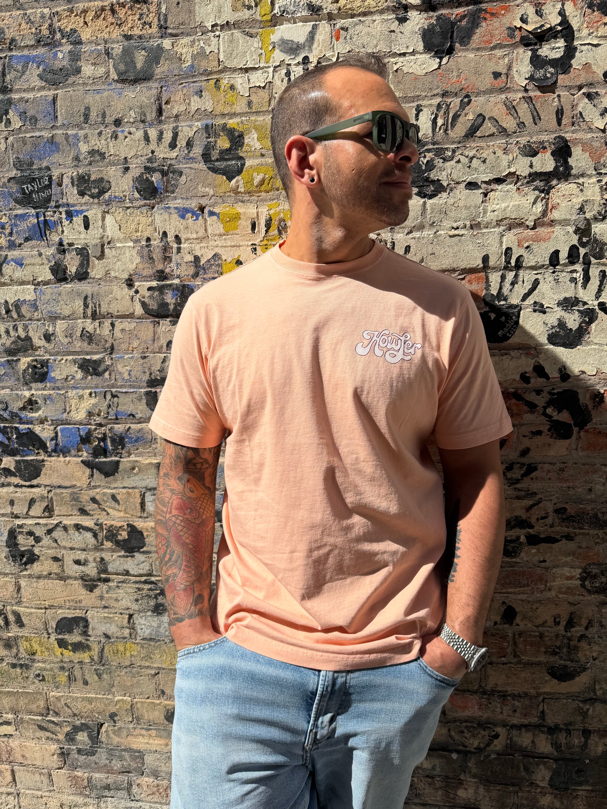 Man wearing a light pink t-shirt with text and blue jeans standing against a textured wall.