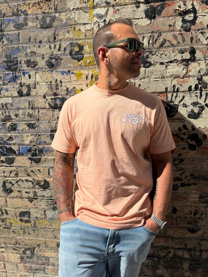 Man wearing a light pink t-shirt with text and blue jeans standing against a textured wall.