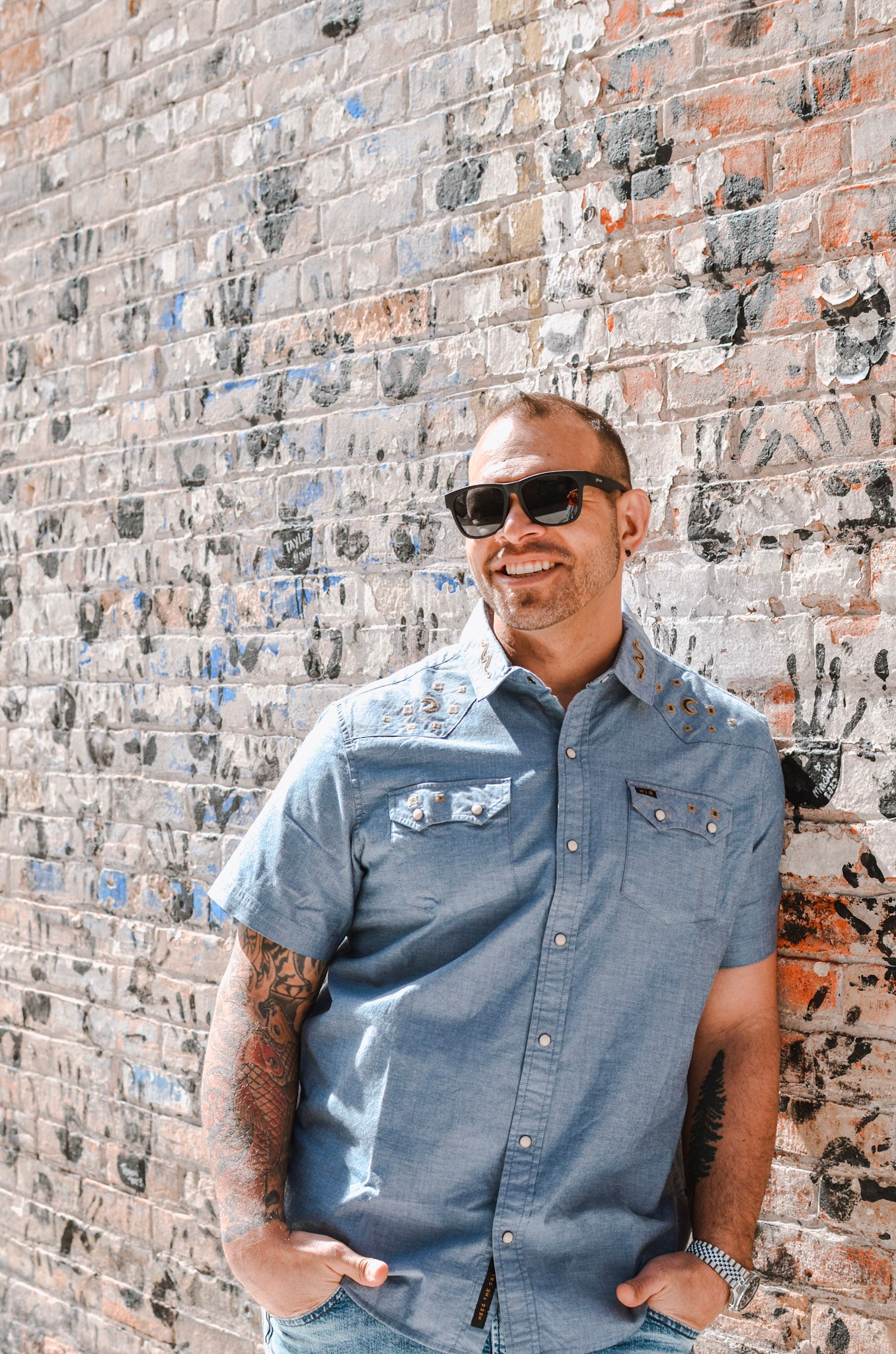 Man wearing sunglasses and a blue shirt standing against a textured brick wall.