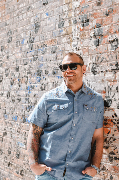 Man wearing sunglasses and a blue shirt standing against a textured brick wall.
