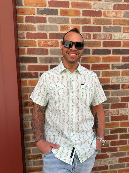 Man wearing sunglasses and a patterned shirt standing against a brick wall.