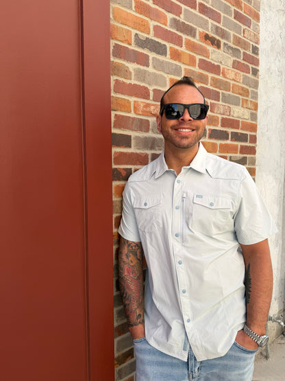 Man wearing sunglasses and a white shirt standing on a brick pavement.