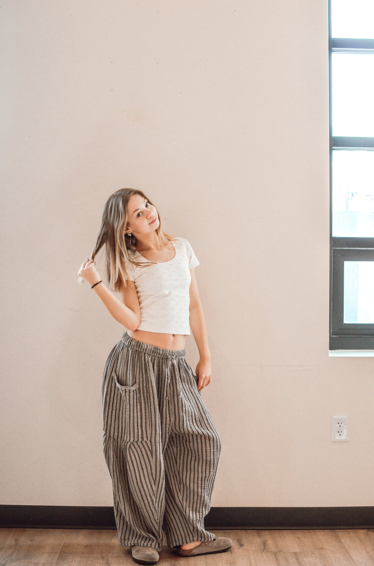 Woman wearing a white top and striped pants standing in a room with a neutral wall and window.