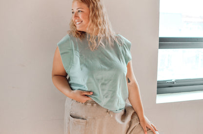 Woman wearing a light green sleeveless top and beige pants standing against a neutral wall.