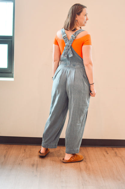 Woman wearing a denim overalls and orange shirt standing in a room with a beige wall.