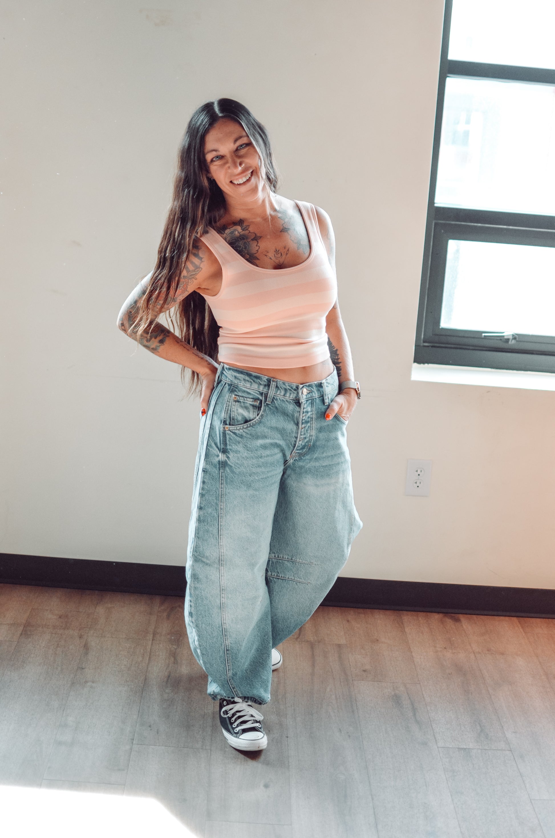 Woman wearing a pink tank top and light blue jeans standing in a room with a window.