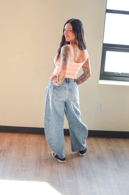 Woman wearing a peach tank top and light blue jeans standing in a room with a window and wall socket.
