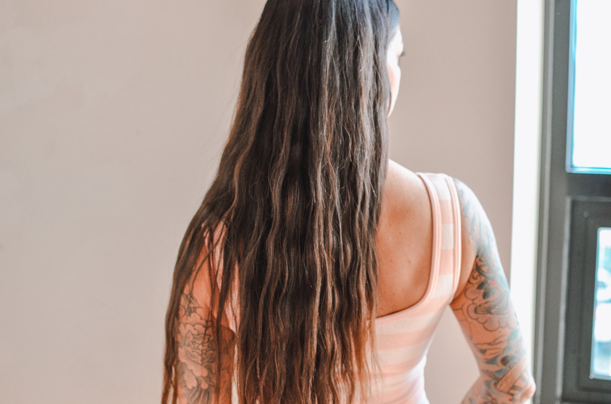 Woman with long hair and tattoos wearing a sleeveless top, standing indoors.
