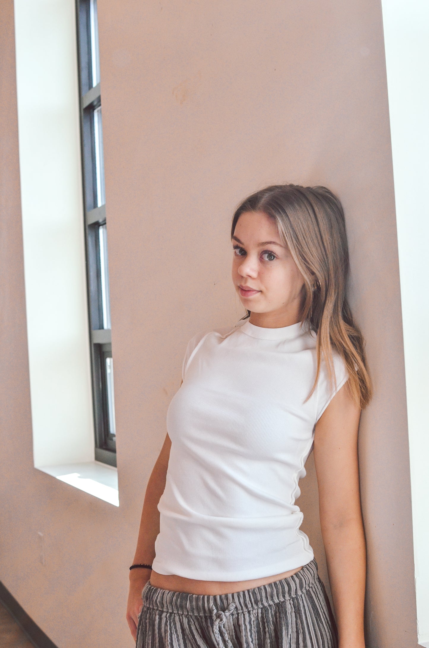 Young woman wearing a white sleeveless top and striped pants against a neutral wall.