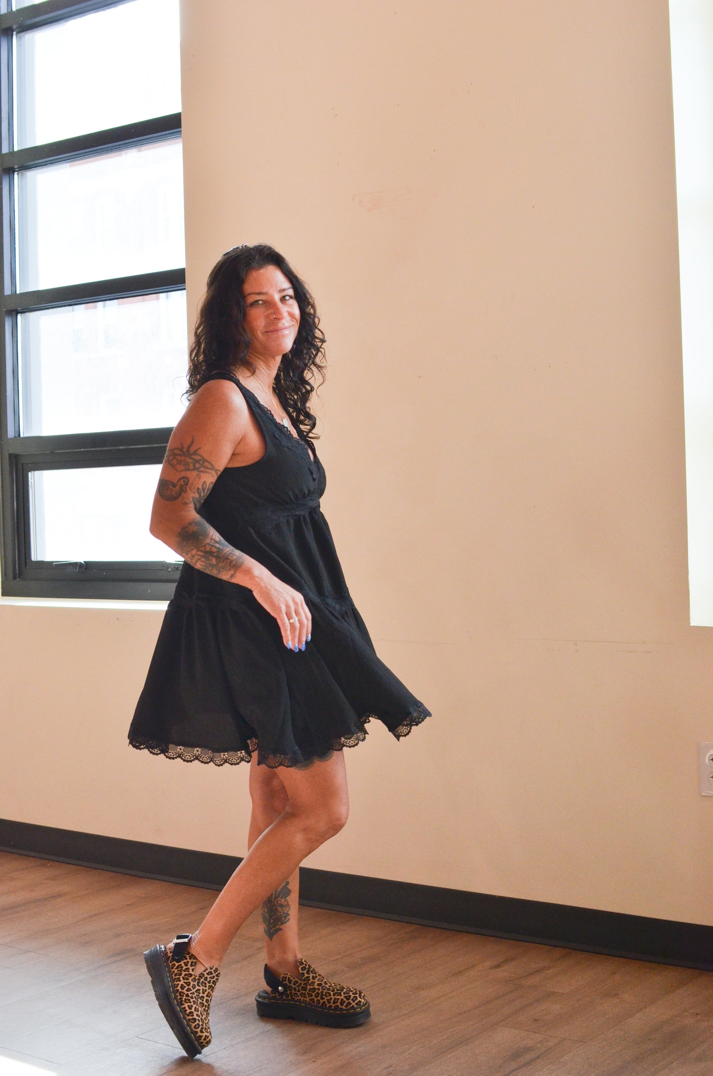 Woman wearing a black dress standing in a room with a window and beige wall.