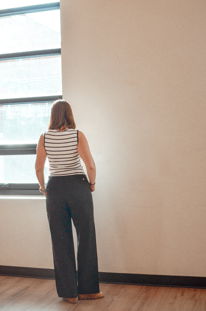 Person wearing a striped shirt and wide-leg pants standing by a window.