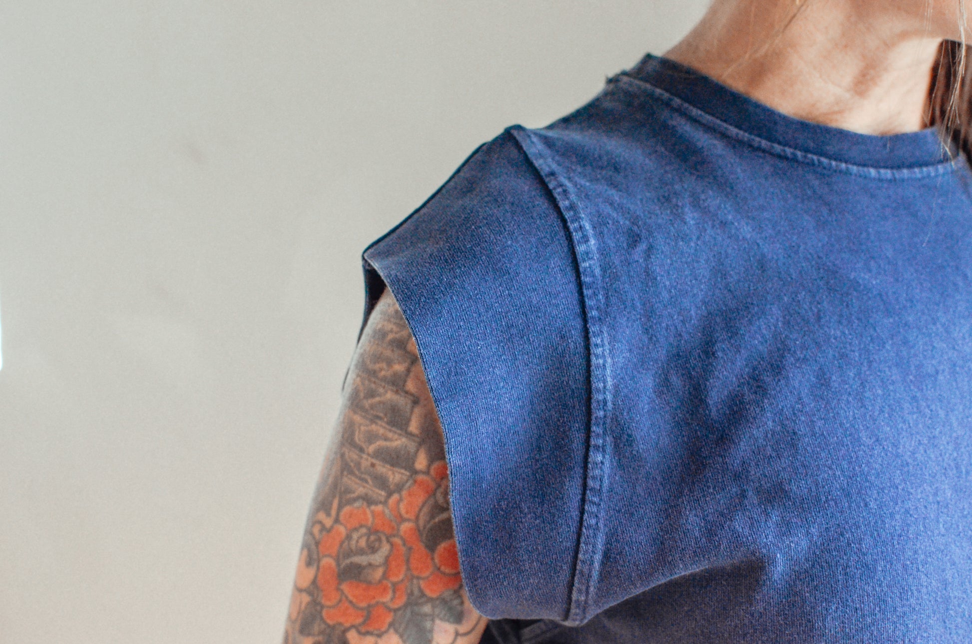 Person wearing a blue sleeveless denim top with a tattooed arm on a plain background