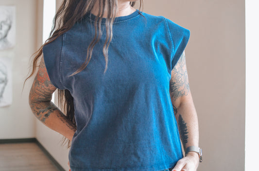 Person wearing a blue sleeveless top with tattoos on arms, standing indoors.