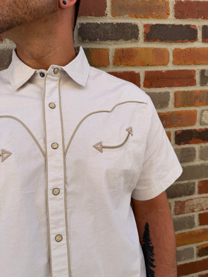 Close-up of a person wearing a white denim jacket with decorative stitching against a brick wall.