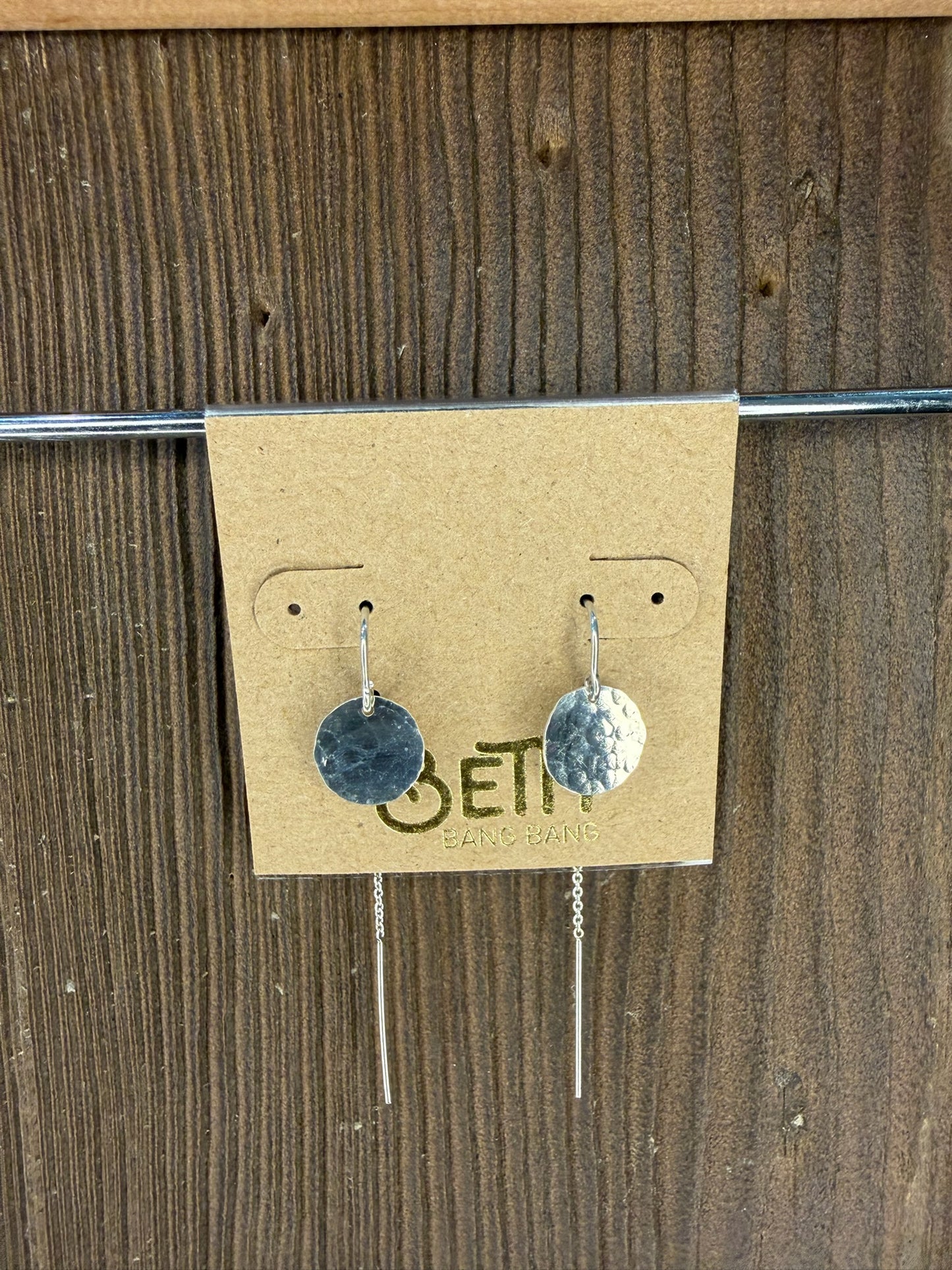 Pair of silver earrings on a cardboard backing with 'Ethereal' branding against a wooden background.
