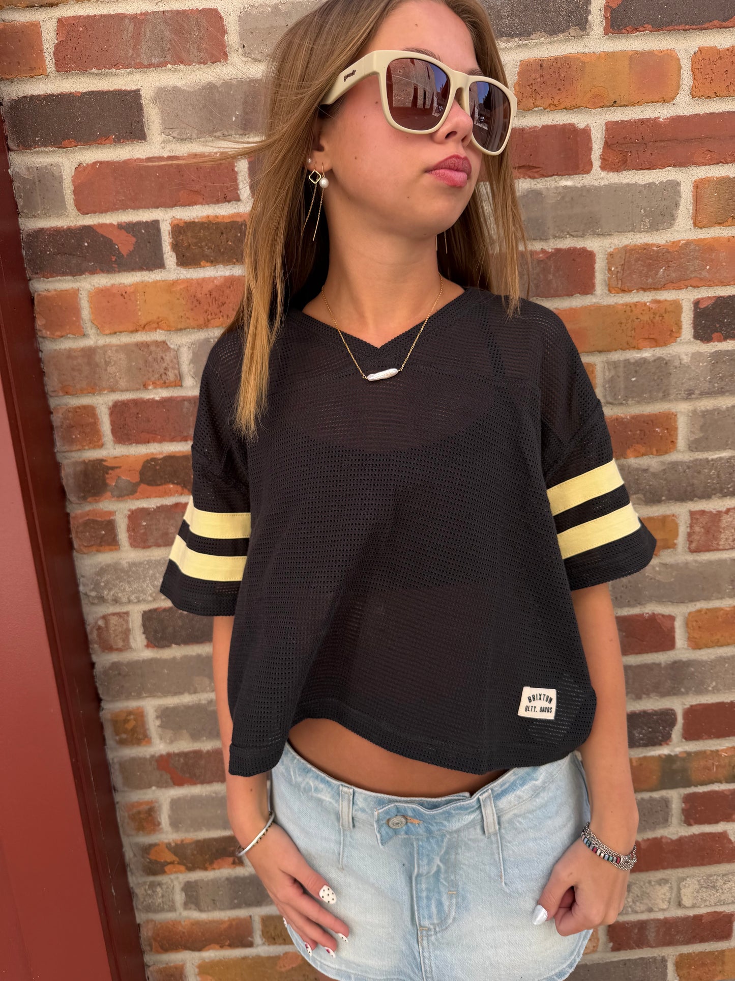 Person wearing a black sweater with yellow stripes and denim shorts against a brick wall.