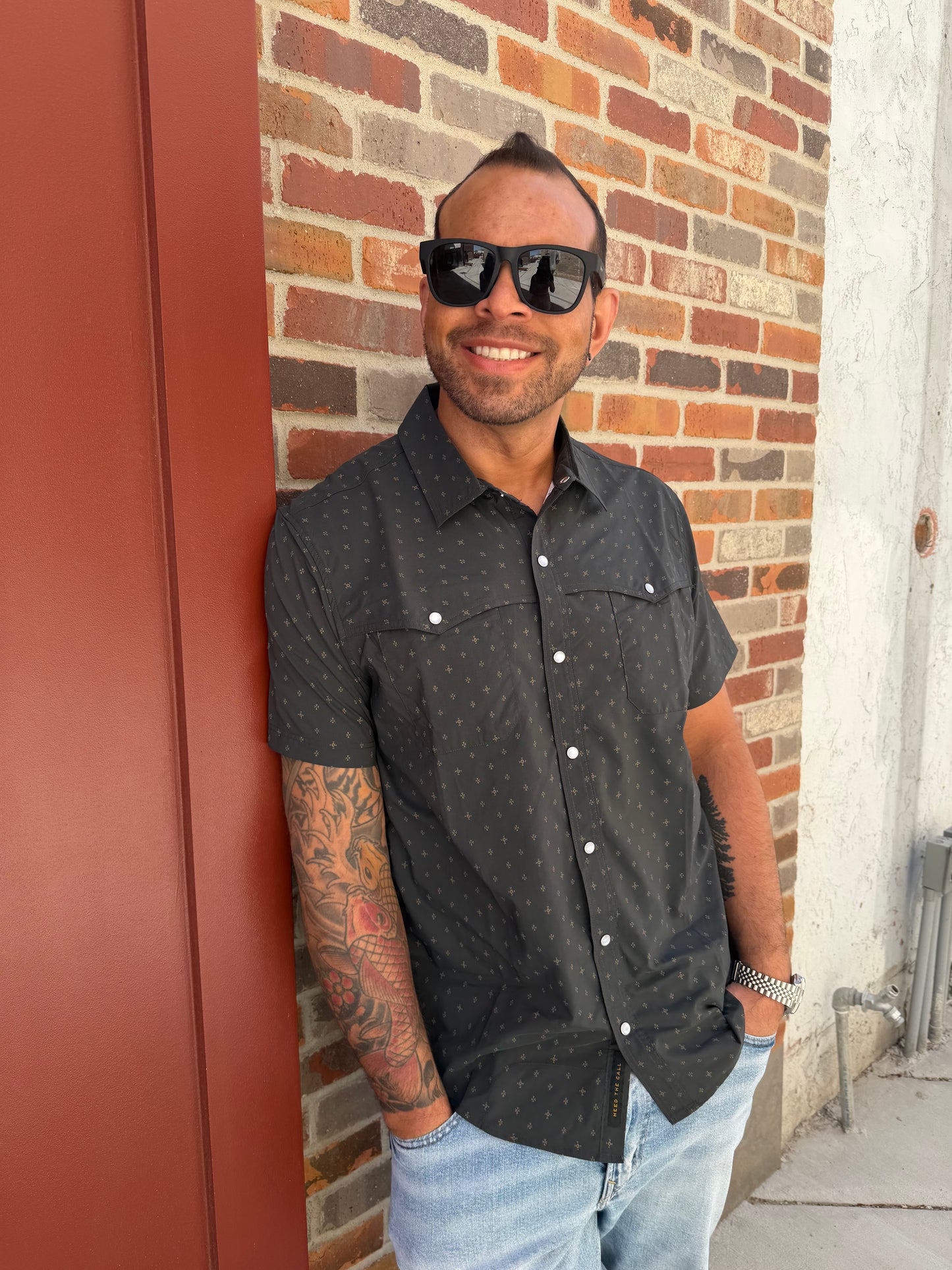 Man wearing a black shirt with white buttons, sunglasses, and jeans standing on a red surface with a brick wall background.