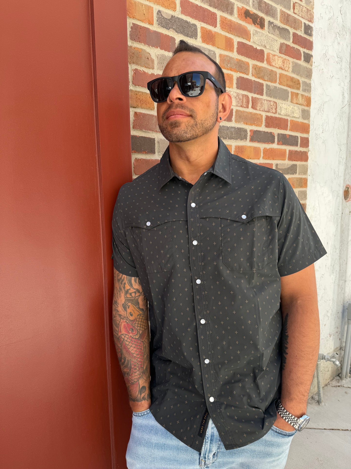 Man wearing a black patterned shirt with white buttons on a red and brown brick pavement.