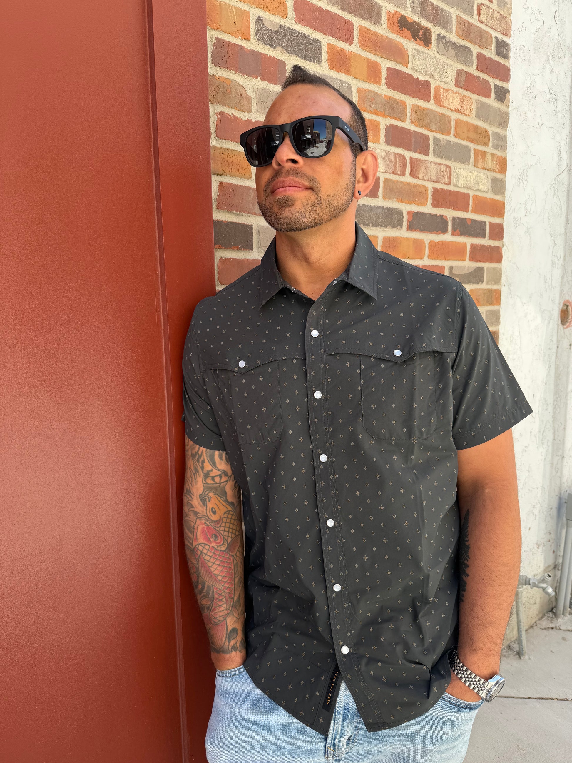 Man wearing a black patterned shirt with white buttons on a red and brown brick pavement.