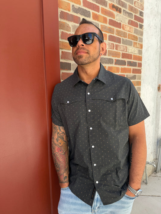 Man wearing a black patterned shirt with white buttons on a red and brown brick pavement.