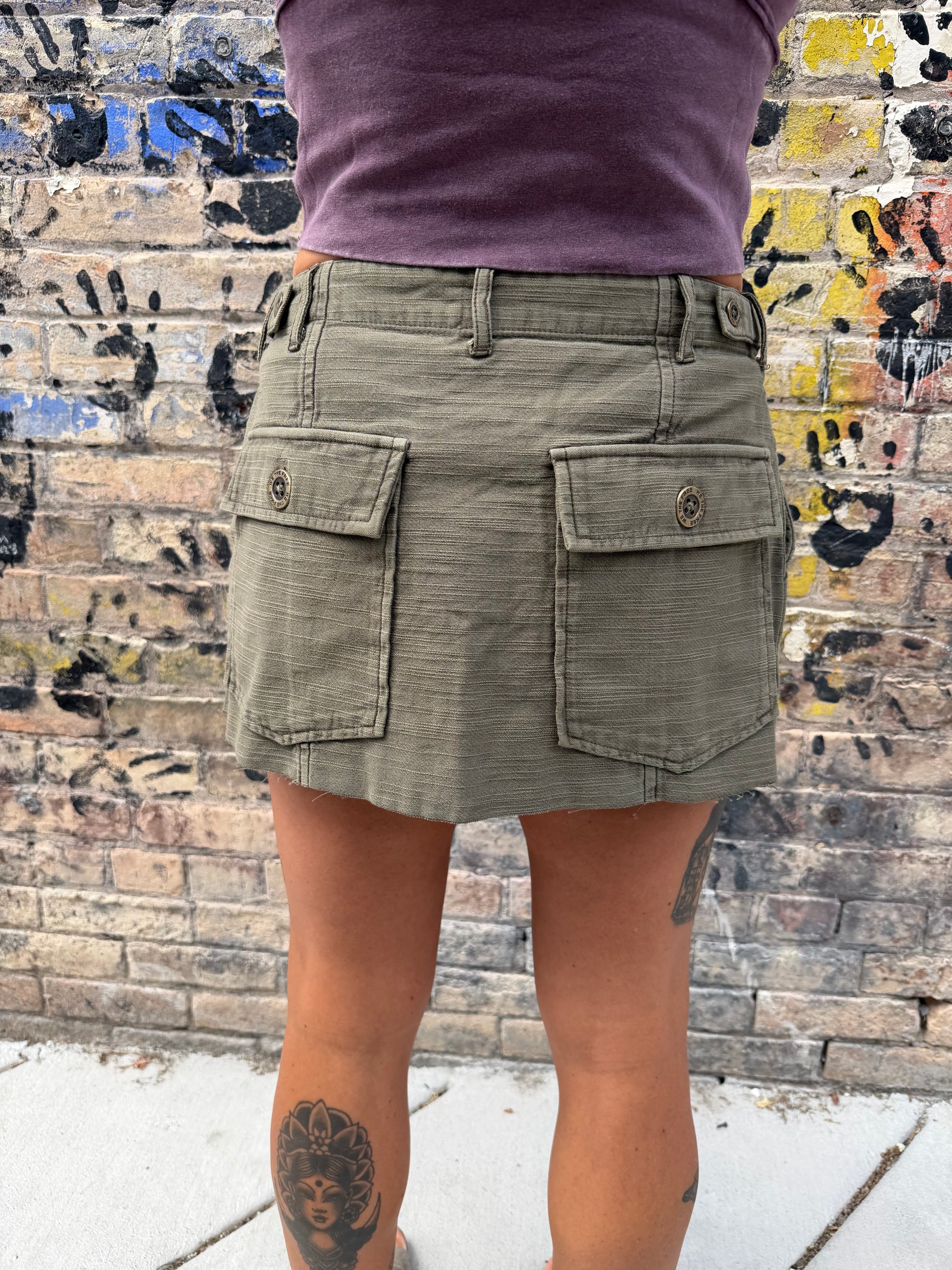 An olive green cargo mini skirt with two large buttoned flap pockets, shown on a model from the back against a graffiti-covered brick wall.