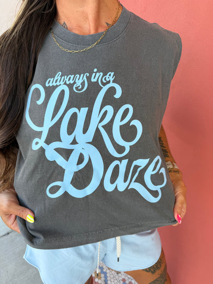 A woman wearing a dark gray sleeveless crop top with light blue script text that reads 'always in a Lake Daze', holding the hem of the shirt against a pink wall.