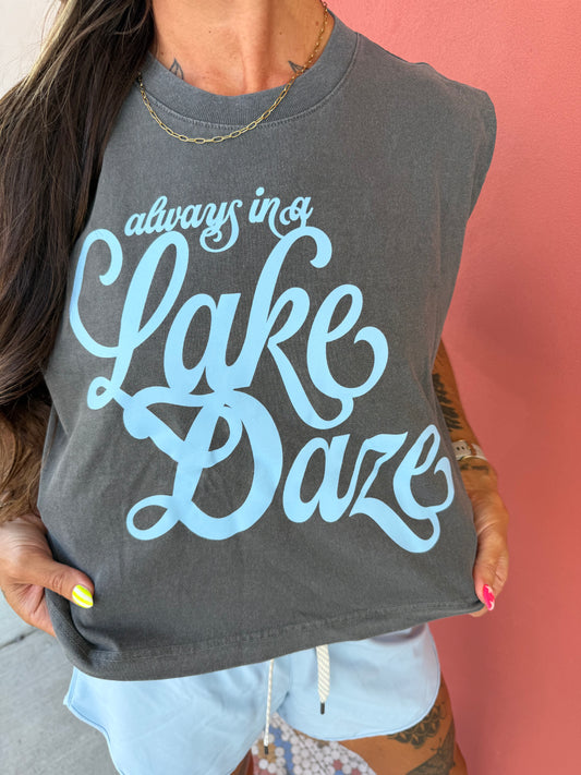 A woman wearing a dark gray sleeveless crop top with light blue script text that reads 'always in a Lake Daze', holding the hem of the shirt against a pink wall.