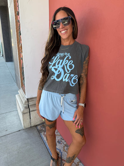 A woman wearing a dark gray graphic t-shirt with light blue text reading 'Always be Lake Daze', paired with light blue shorts, standing against a coral-pink wall.