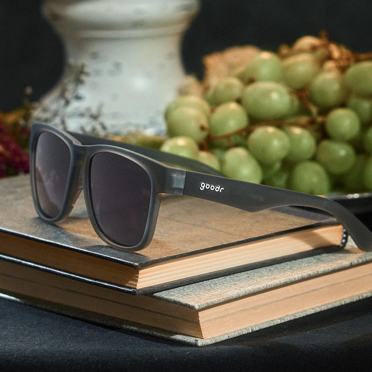Black sunglasses with 'goodr' logo on a stack of books with grapes in the background