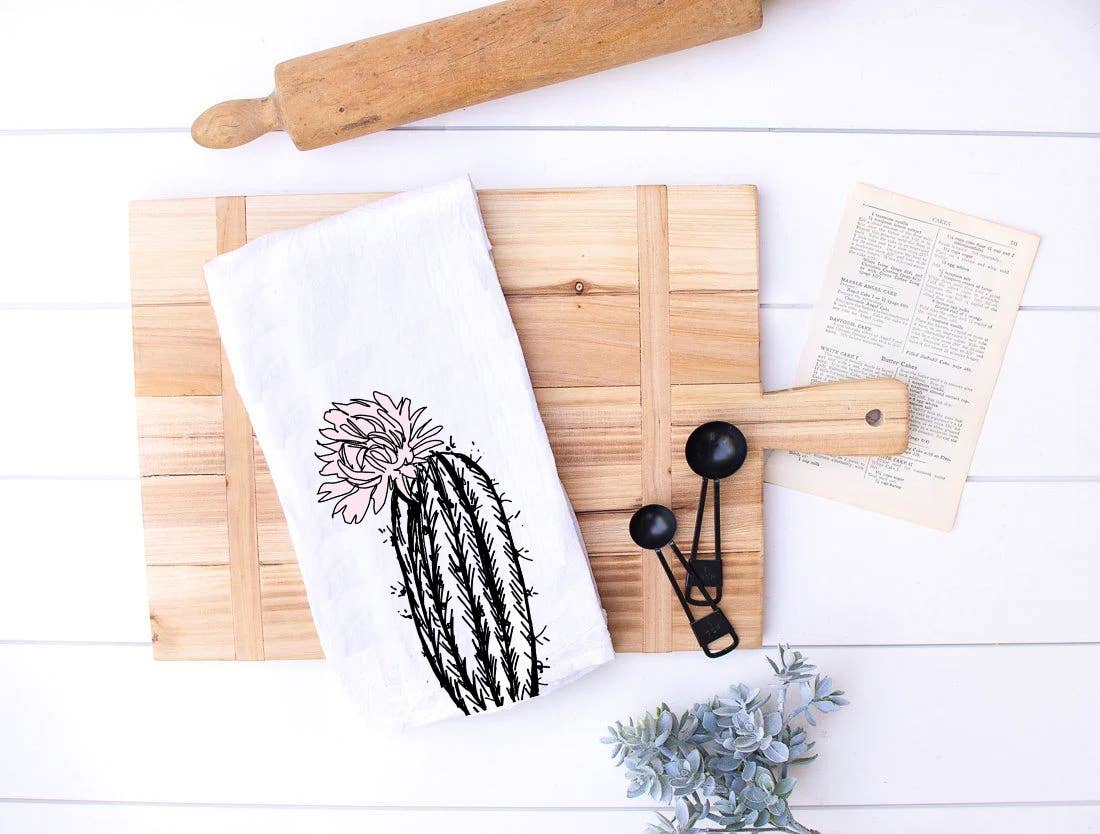 Wooden cutting board with a cactus-themed kitchen towel, rolling pin, and small plant on a white surface.