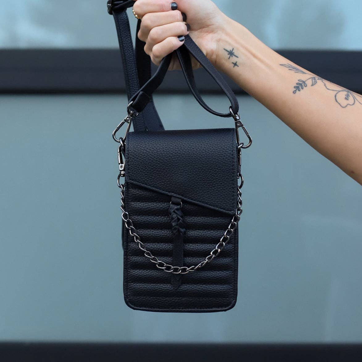 Black leather crossbody bag with chain detail held by a hand against a neutral background