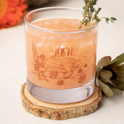 Cocktail in a glass with a desert-themed engraving on a wooden coaster.