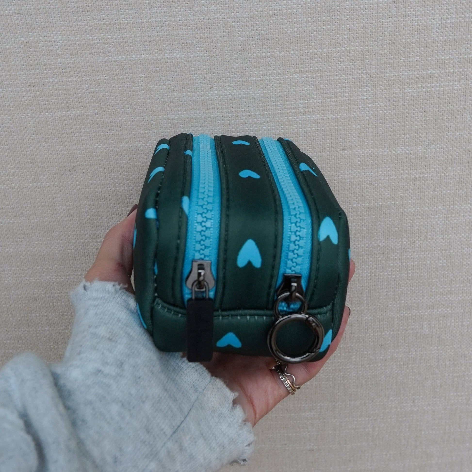 Hand holding a small green pouch with blue zippers and heart patterns against a beige background