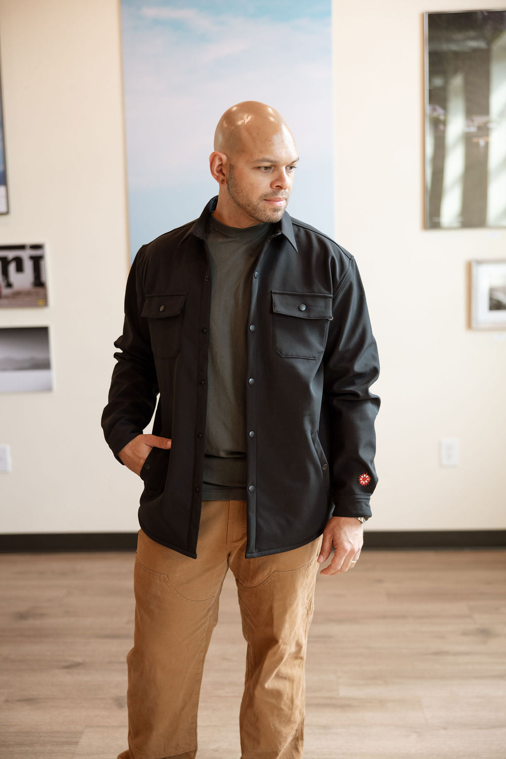 Man wearing a black jacket and brown pants standing in an indoor setting with a neutral background.