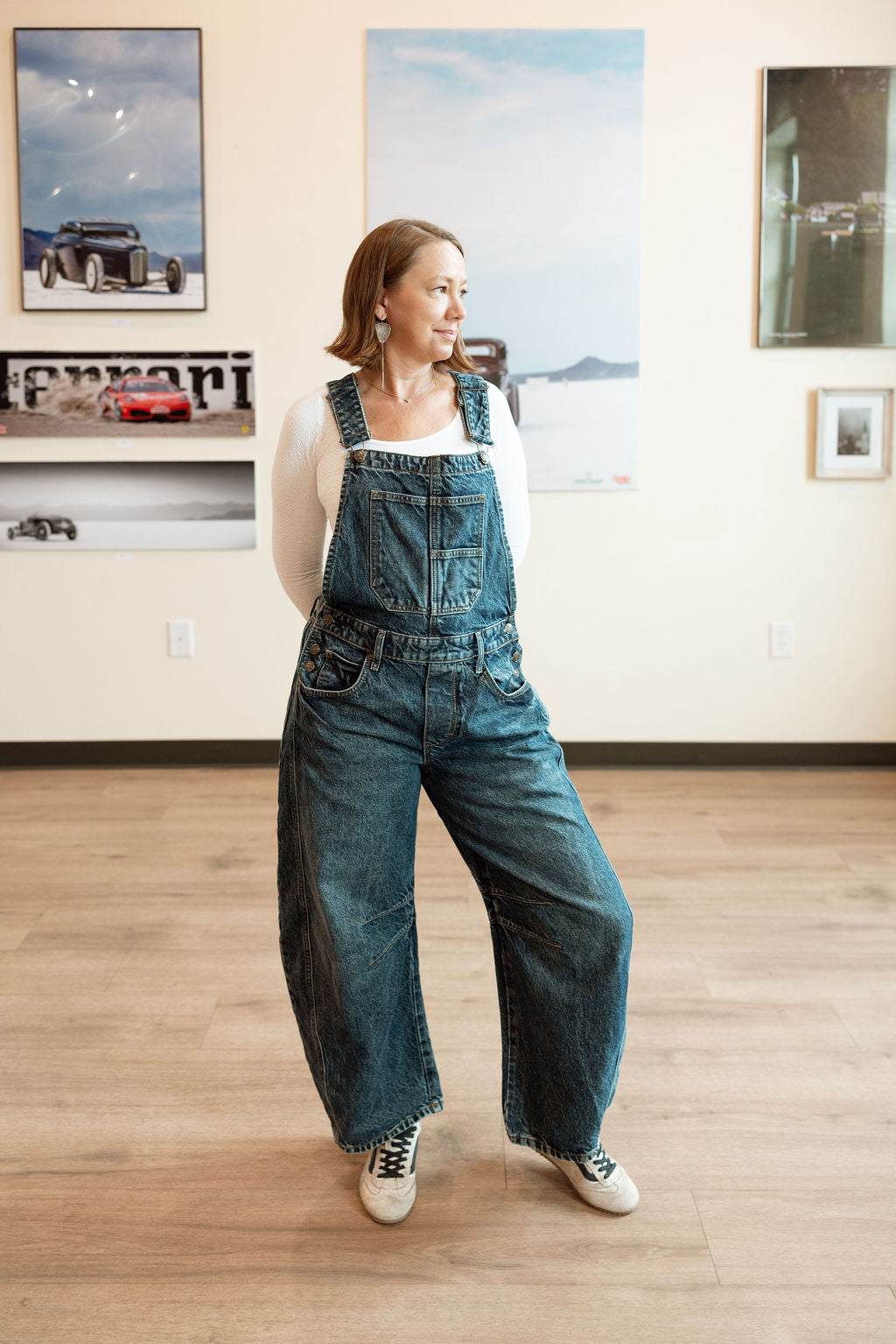 Woman wearing blue denim overalls standing in a room with framed pictures on the wall.